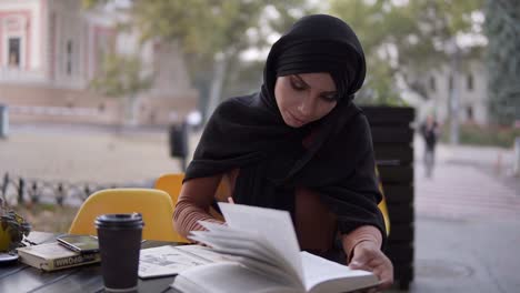 an attractive muslim girl writes in a notebook sitting in a cafe with a book. studying in outdoors cafe, having break. college, self education. front view