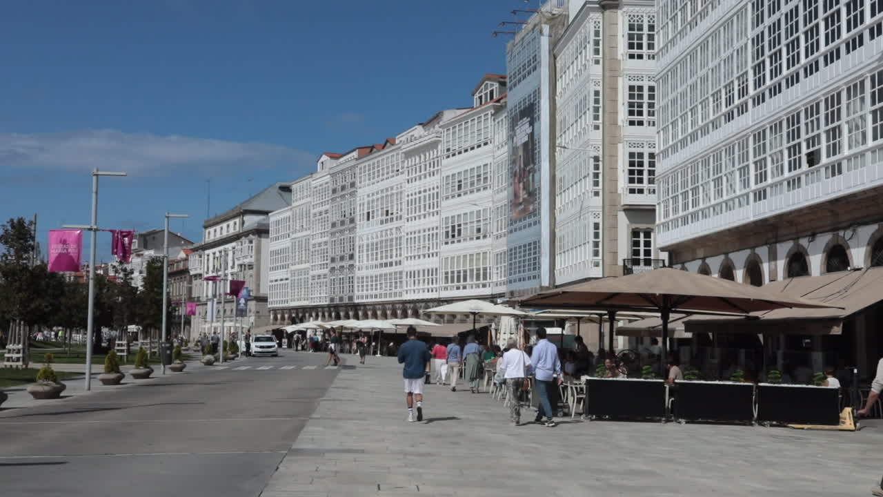 Pedestrians stroll past cafés on a sunny day in front of white glass-fronted buildings