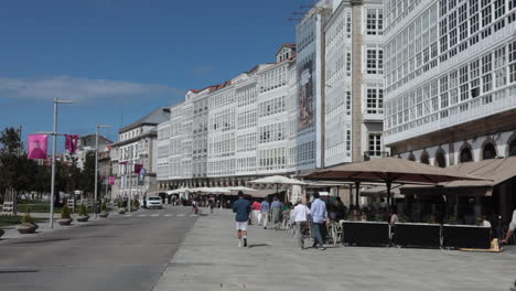 pedestrians stroll past cafés on a sunny day in front of white glass-fronted buildings