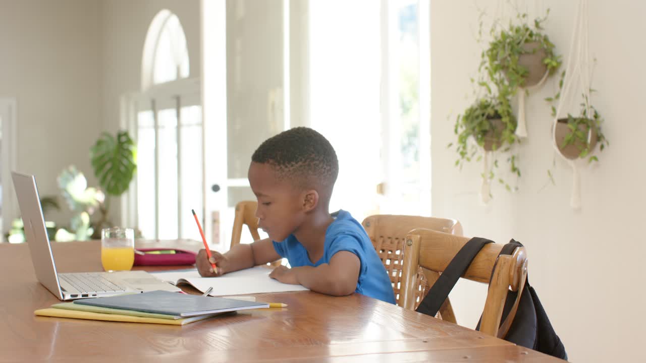 African American child studying with red pencil at dining table as parent coming adjusting backpack