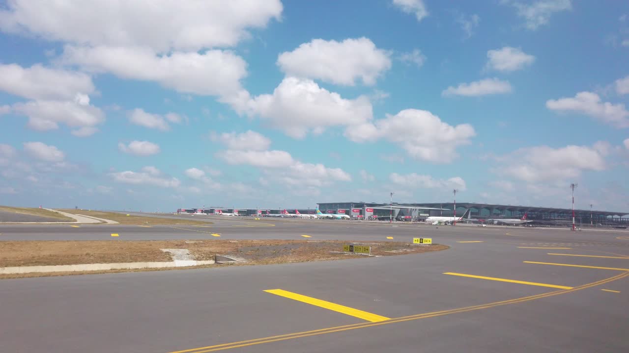 On a partly cloudy daytime, the view from the airplane window captures Istanbul Airport's runways, with the terminal building in the background and parked airplanes scattered around
