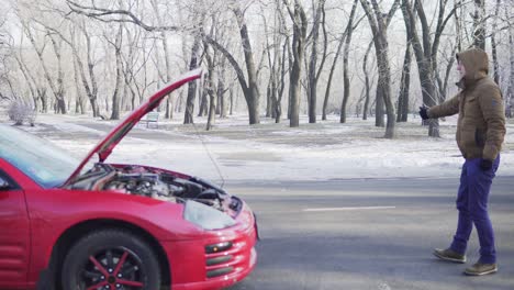 un joven atractivo haciendo autostop en una carretera con su coche averiado cerca de él