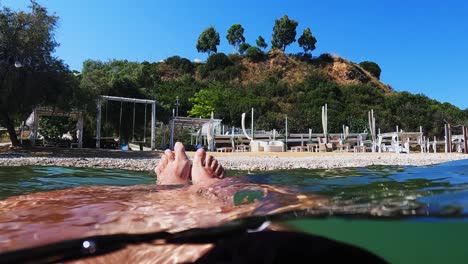 Personal-perspective-of-male-legs-and-feet-relaxing-while-floating-on-sea-water-with-swing-on-beach-in-background