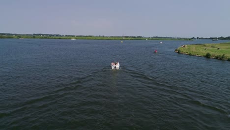 Drone-Slomo-shot-of-Small-Boat-with-5-Male-Friends-Sailing-surrounded-by-Dutch-Countryside-during-Sunny-weather-with-Windmill-in-background