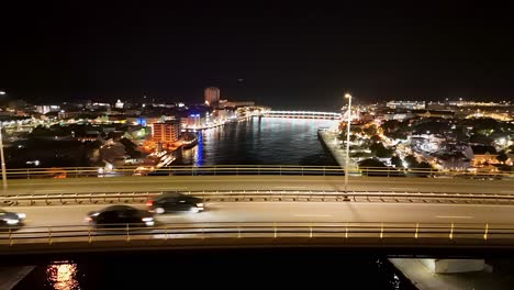 Seguimiento-Aéreo-Panorámico-A-Través-Del-Puente-Reina-Juliana-Mientras-Los-Autos-Pasan-A-Toda-Velocidad-Por-La-Noche,-Handelskade-Willemstad-Curacao-Al-Fondo