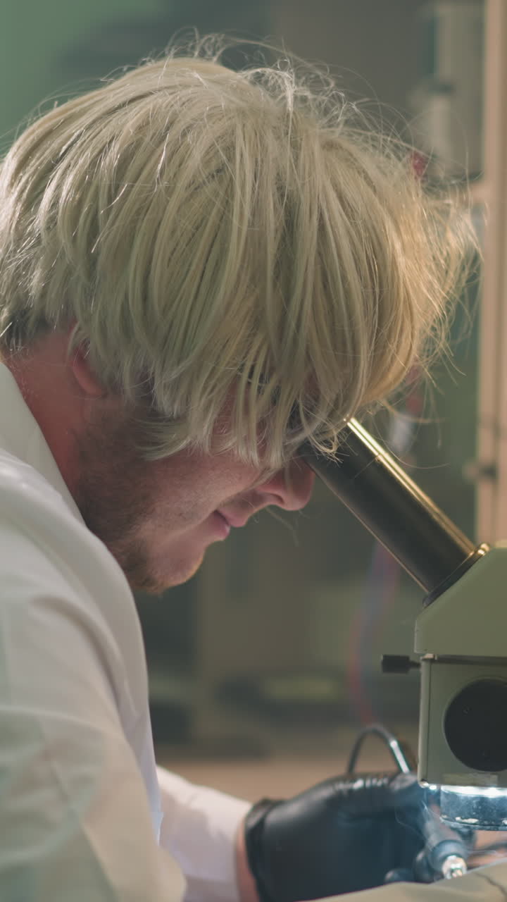 una vista de cerca de un técnico con una peluca rubia intensamente enfocado en un microscopio en un entorno de laboratorio