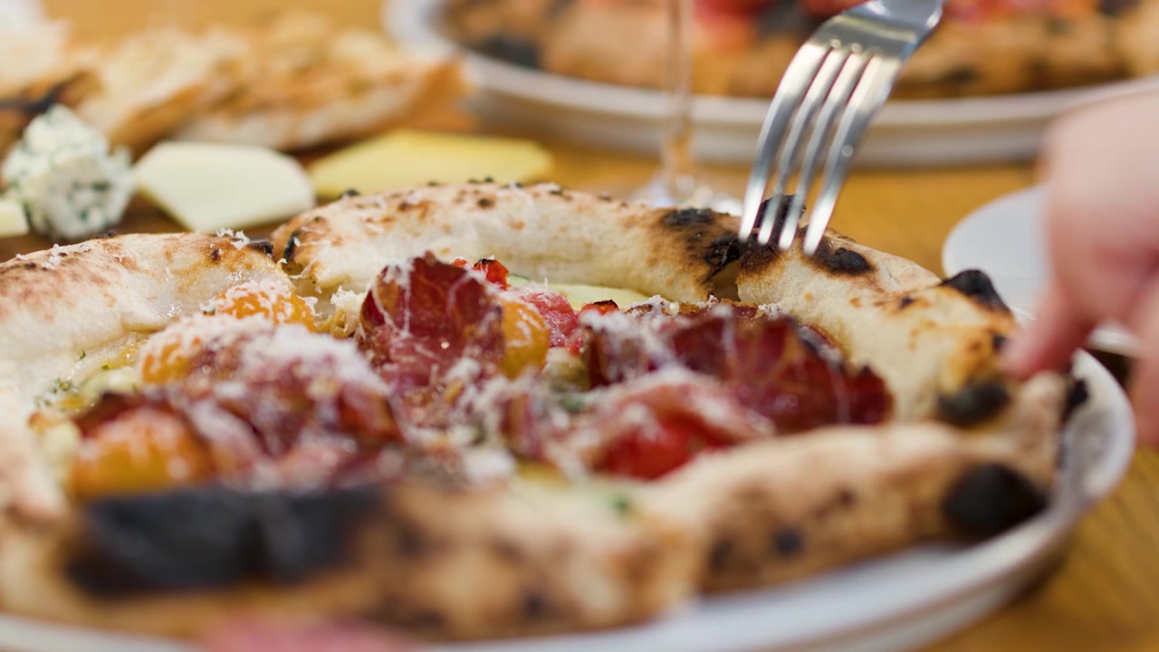 Caucasian hands use a fork to eat gourmet Neapolitan pizza at a restaurant table.