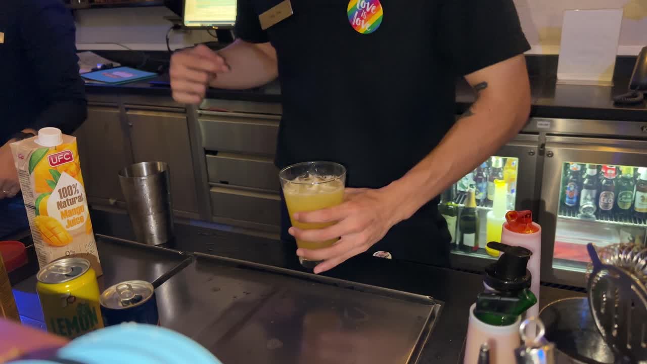 Bartender in black uniform prepares a fresh juice beverage behind a modern bar counter under artificial lighting, using cartons, shakers, and glassware