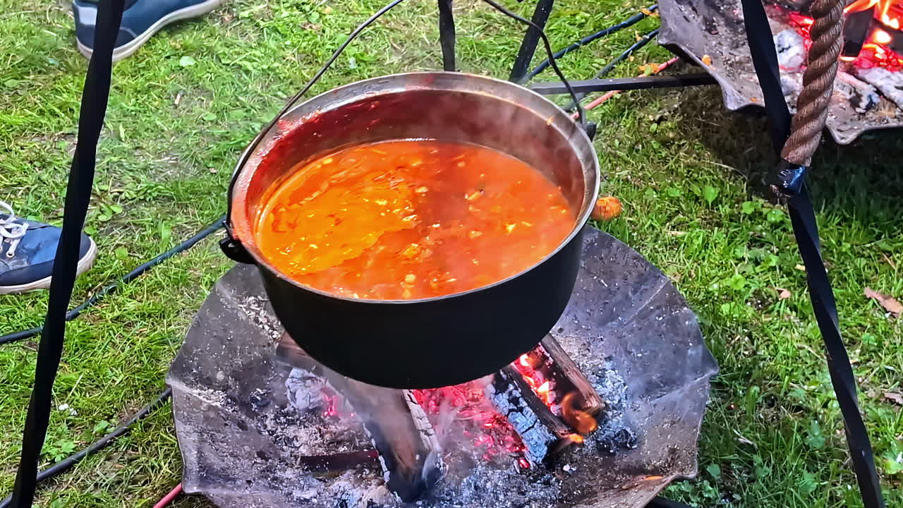 Orange soup bubbles in metal pot over open fire suspended by tripod during outdoor cooking