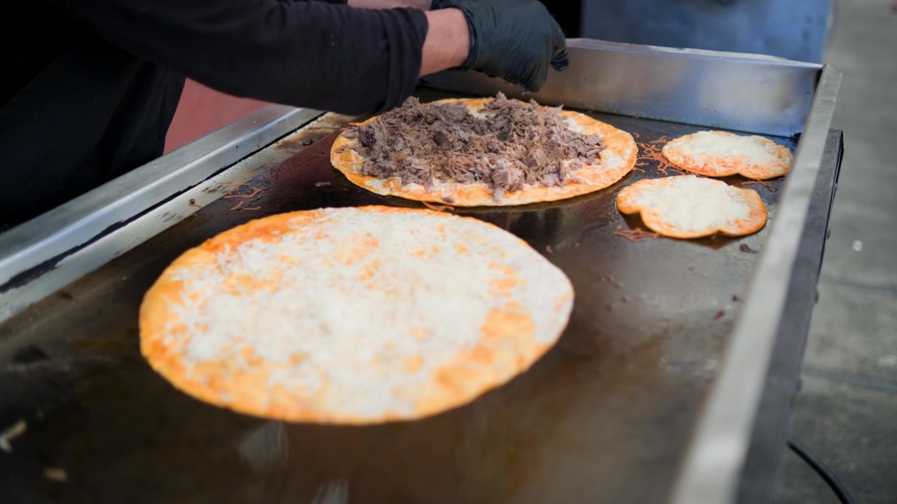 Commercial kitchen footage showing adding birria meat to the birria pizza during preparation of a savory birria pizza on a flat grill
