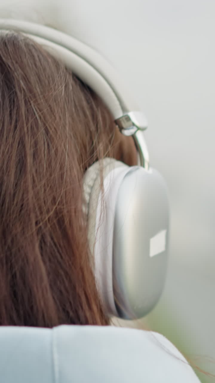 Rear view of student with brown hair wearing white headphones, strolling outdoors into blurred background, serene atmosphere, youthful and focused, peaceful walk in urban environment