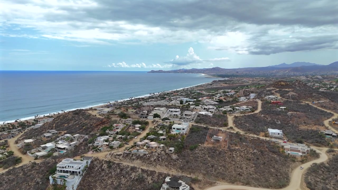 Drone se move para a esquerda sobre a comunidade na encosta de La Laguna, voando alto o suficiente para ver o oceano e a praia abaixo, com céus claros e Palmilla Point visível à distância, enquanto nuvens mais escuras se formam.