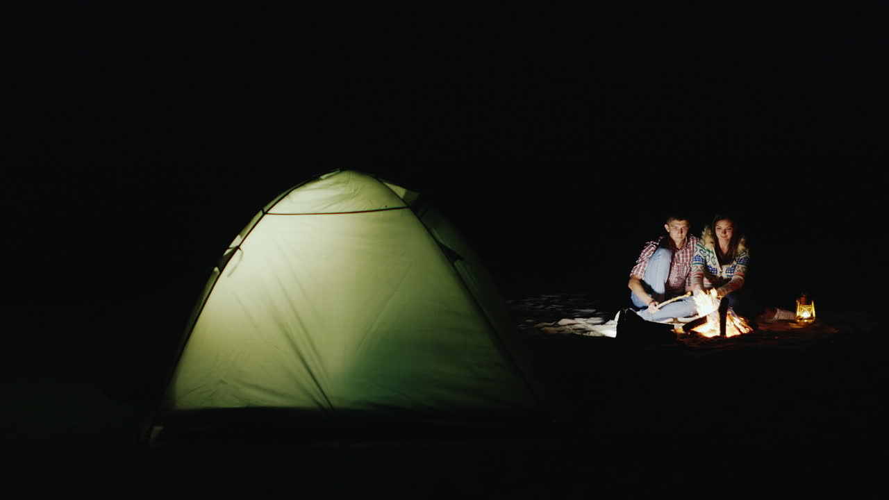 젊은 부부는 모닥불과 텐트 주변의 막대기에 마시멜로를 굽습니다.
