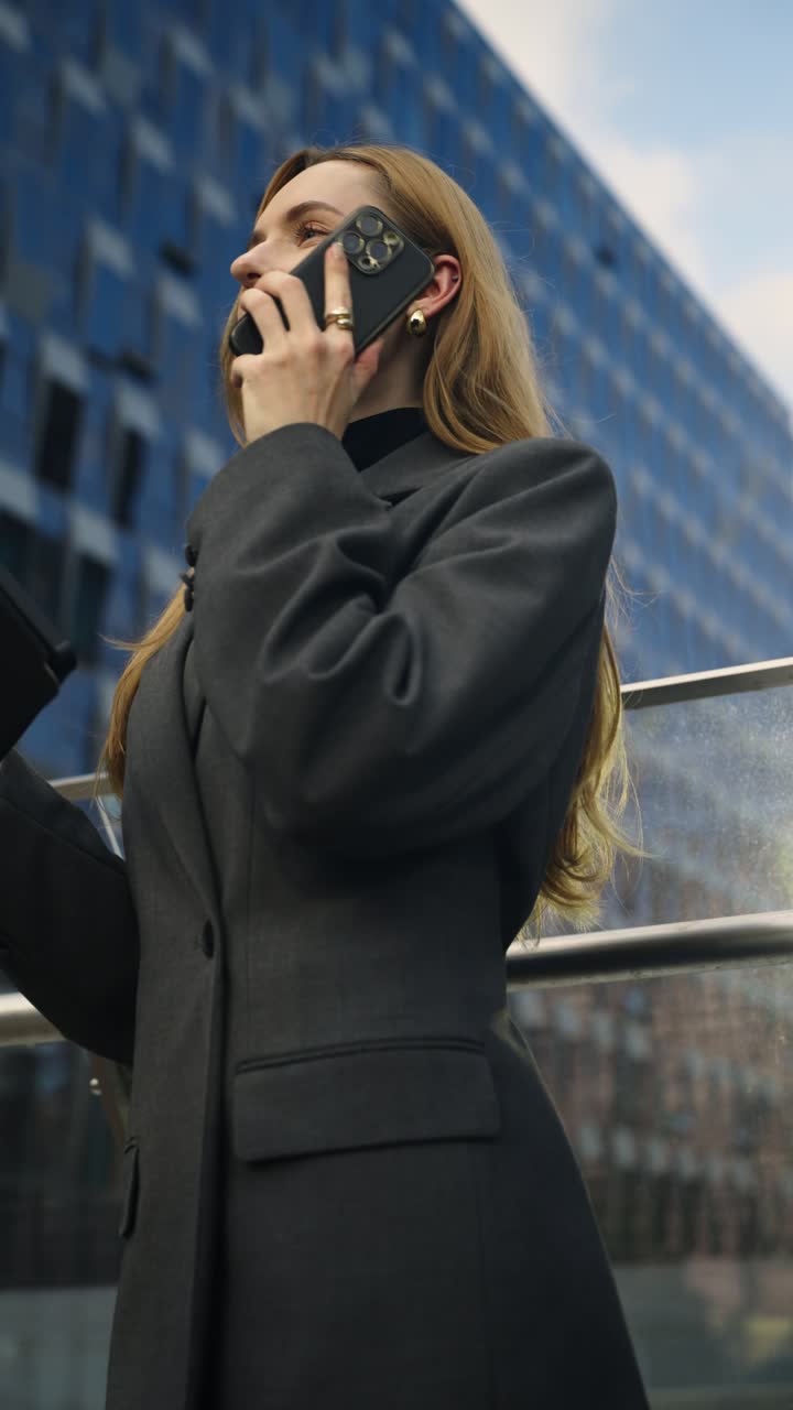 Young businesswoman in a gray blazer talks on her phone while checking a digital tablet outdoors by glass offices