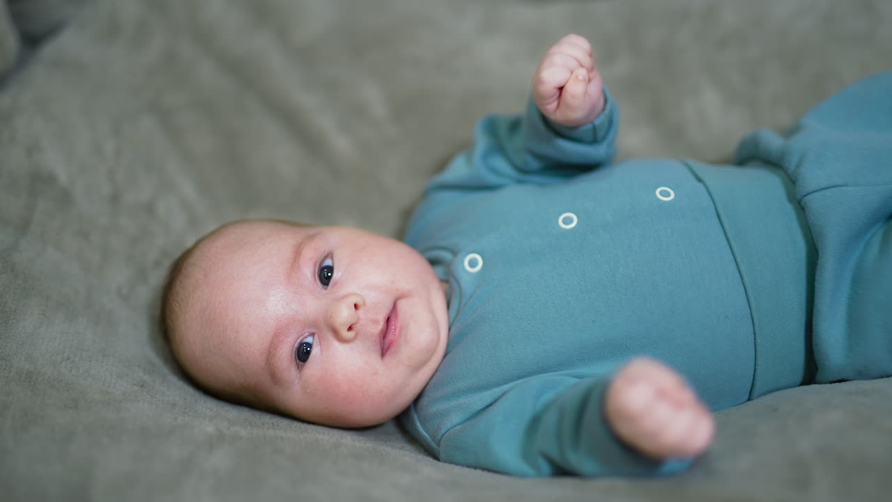 Funny baby on the bed with his head turned right. Beautiful baby looks into camera and sneezes. Gray backdrop.
