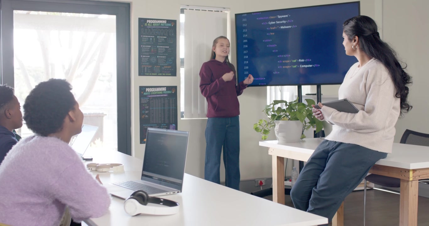 Teen African female starting presentation, pointing to code on monitor in lab, teaching peers