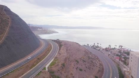 Blick-Von-Einer-Drohne,-Die-über-Eine-Autobahn-Zurückfliegt,-Mit-Einem-Berg-Auf-Der-Linken-Seite-Und-Der-Hotelzone-Auf-Der-Rechten-Seite-Nahe-Der-Küste,-Die-Das-Meer-Und-Den-Himmel-Im-Hintergrund-In-Mexiko-Zeigt