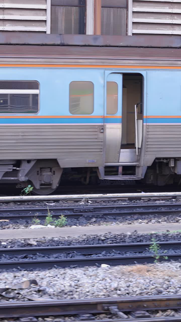 Tampilan samping lokomotif diesel berwarna cerah yang menarik gerbong penumpang biru melalui stasiun Bangkok.