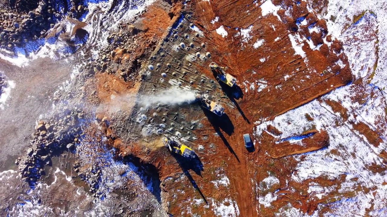 Quarry drilling rig operation, Bulgaria. Top down drone shot of three rigs boring blast holes in rust colored bedrock with dust rising.