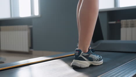 lower angle view of person walking on treadmill with sunlight shining around leg and sneakers under bright modern indoor gym light, exercise bikes and elliptical trainer blurred behind on tiled floor