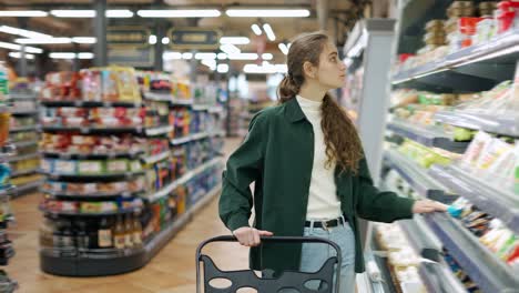 Una-Mujer-En-Un-Supermercado-Empujando-Un-Carrito,-Eligiendo-Queso-En-La-Sección-De-Leche.