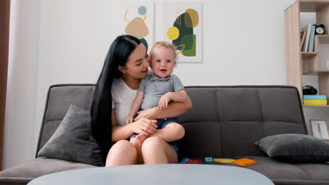 Beautiful-Mom-Sitting-On-Sofa-In-Living-Room-Holding-Her-Adorable-Little-Boy-And-Looking-At-Camera-While-Having-A-Video-Call