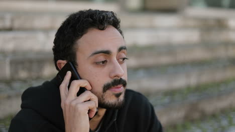 Closeup-of-young-Arabic-mans-face-with-dark-curly-hair-and-beard-in-black-hoodie-sitting-on-stairs-outside