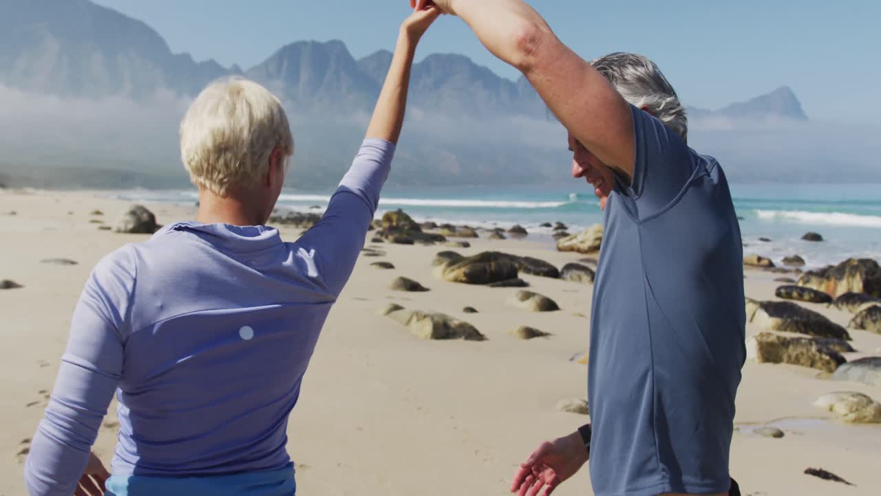 Pareja de senderistas mayores bailando en la playa mientras hacen senderismo.