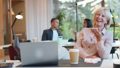 Mature-Businesswoman-With-Feet-On-Desk-Working-In-Office-Talking-Into-Mic-Of-Mobile-Phone