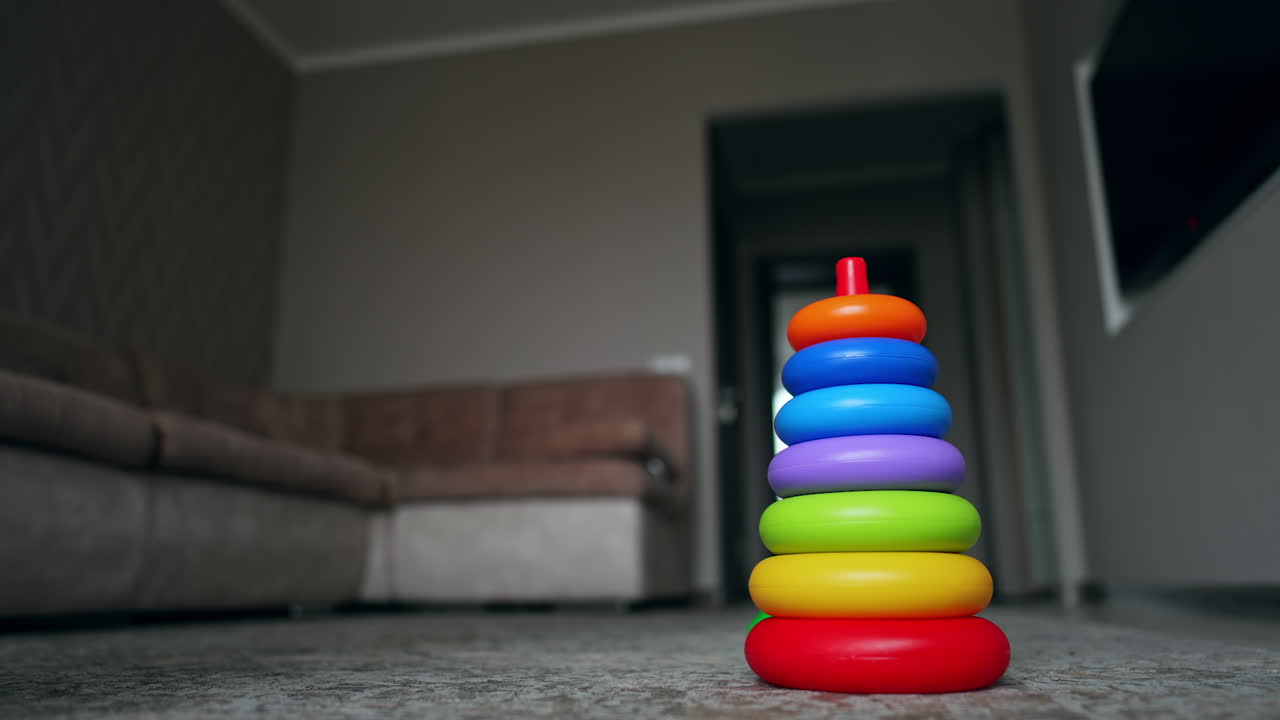 Pyramid without a top stands on the floor. Unrecognized baby in checkered pants jumps up to a toy and puts red ring. Low angle view.