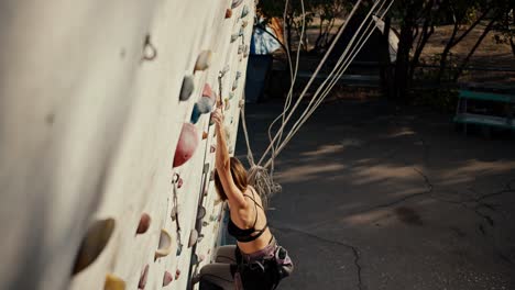 vista superior de una chica rubia con un peinado bob en una camiseta negra y pantalones negros. ella sube a una estructura especial para escaladores y trenes. una chica en equipo de escalada sube a unha estructura especial para entrenamiento de escalada durante el día