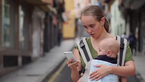 Una-Joven-Madre-Está-De-Viaje-Con-Su-Bebé-Asegurado-En-Una-Mochila-Canguro.-Utiliza-Su-Teléfono-Móvil-Para-Tomar-Fotografías-Y-De-Vez-En-Cuando-Mira-La-Pantalla-Mientras-Camina