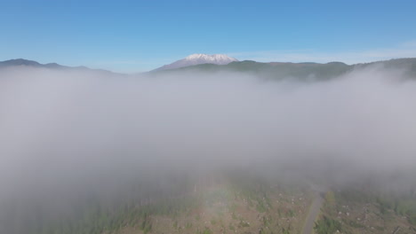 Drone-dolly-over-empty-road-to-thick-blanket-of-fog-shrouding-surrounding-valley,-Mount-St-Helens-on-horizon