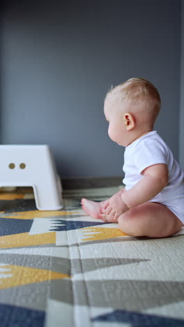 beautiful plump blond baby in white bodysuit crawling by the floor. child reaches the chair and tries to stand up. vertical video.