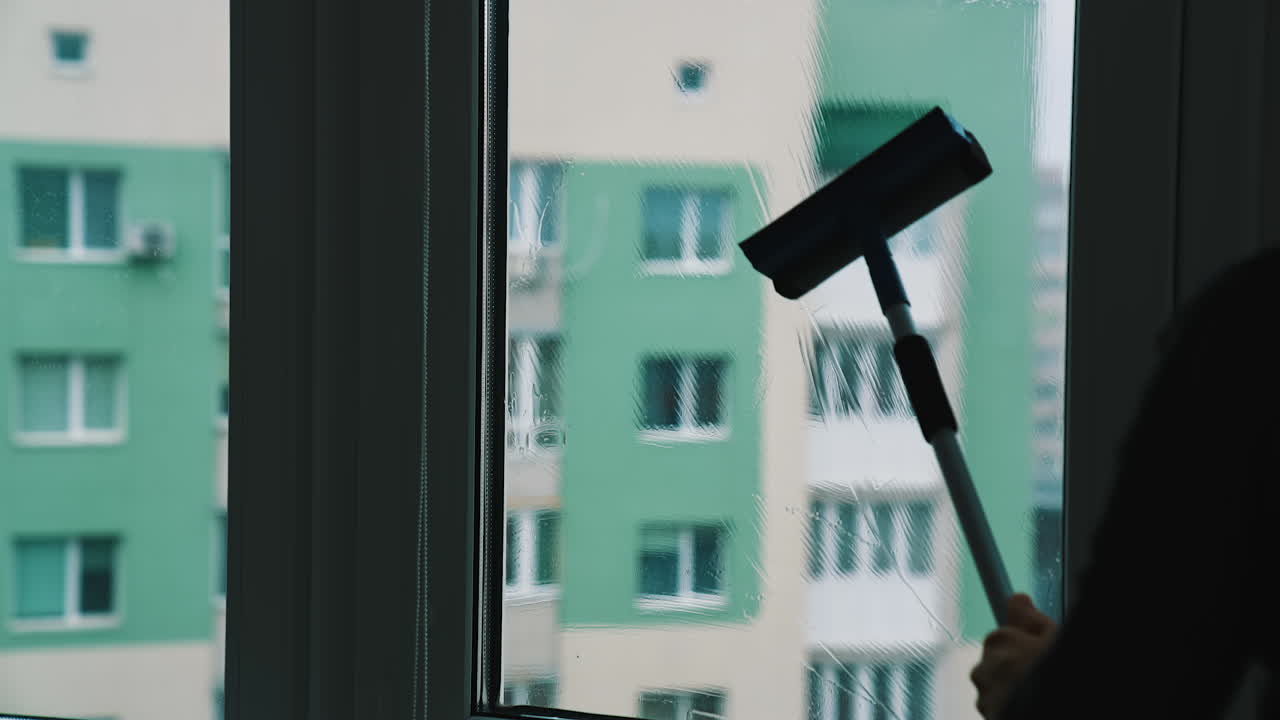 Process of washing window at home in the city. Silhouette of a man cleaning glass window with a brush and special liquid in flat on high building background.