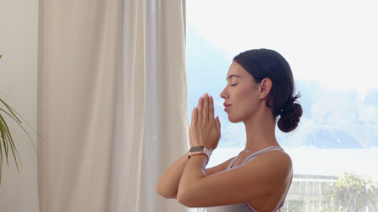 Woman practicing yoga at home, meditating peacefully by window in morning light, copy space