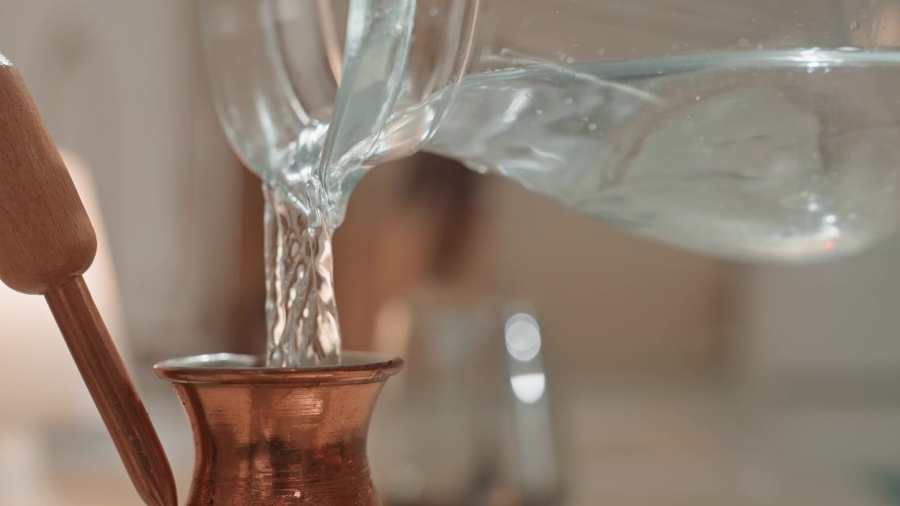 Primo piano di una cezve di rame che riceve un versamento delicato, flusso misurato e morbidi riflessi sul metallo, composizione da tavolo con sfondo sfocato, barista casalingo e appassionato di artigianato che crea un ritmo ASMR.