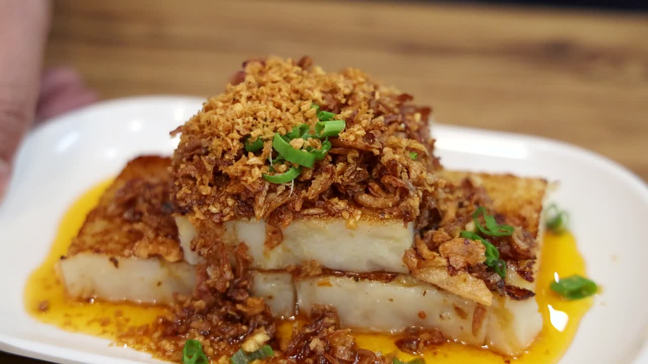 A plate of fried turnip cake topped with crispy shallots and green garnish is presented on a wooden table under warm, natural lighting with smooth camera movement