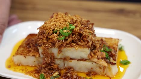 a plate of fried turnip cake topped with crispy shallots and green garnish is presented on a wooden table under warm, natural lighting with smooth camera movement