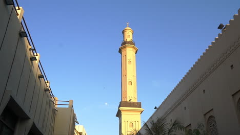 view of the tower of grand bur dubai masjid mosque with blue sky on the background in deira gold souk, dubai, uae from the alley. -wide shot