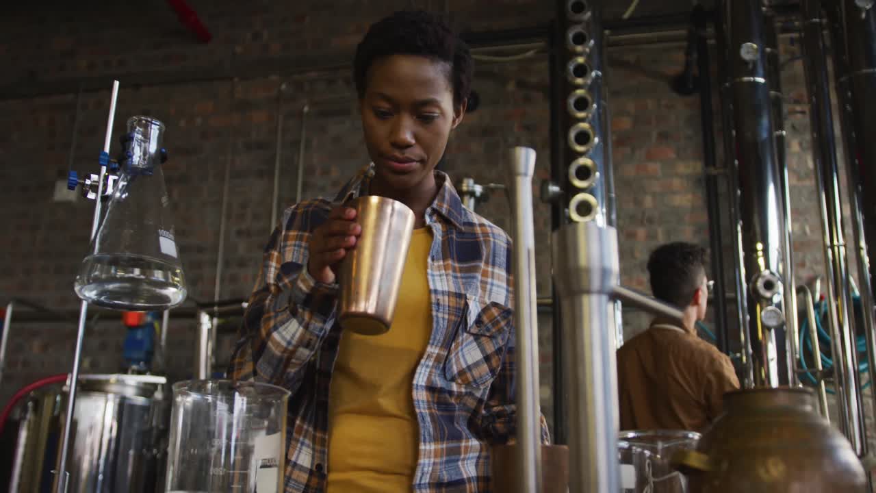 African american woman working at gin distillery smelling product and using tablet