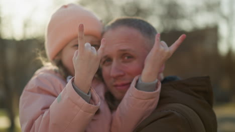 a man wearing a brown jacket carries a little girl in a pink cap and jacket as she makes a playful hand sign with both hands and they smile joyfully