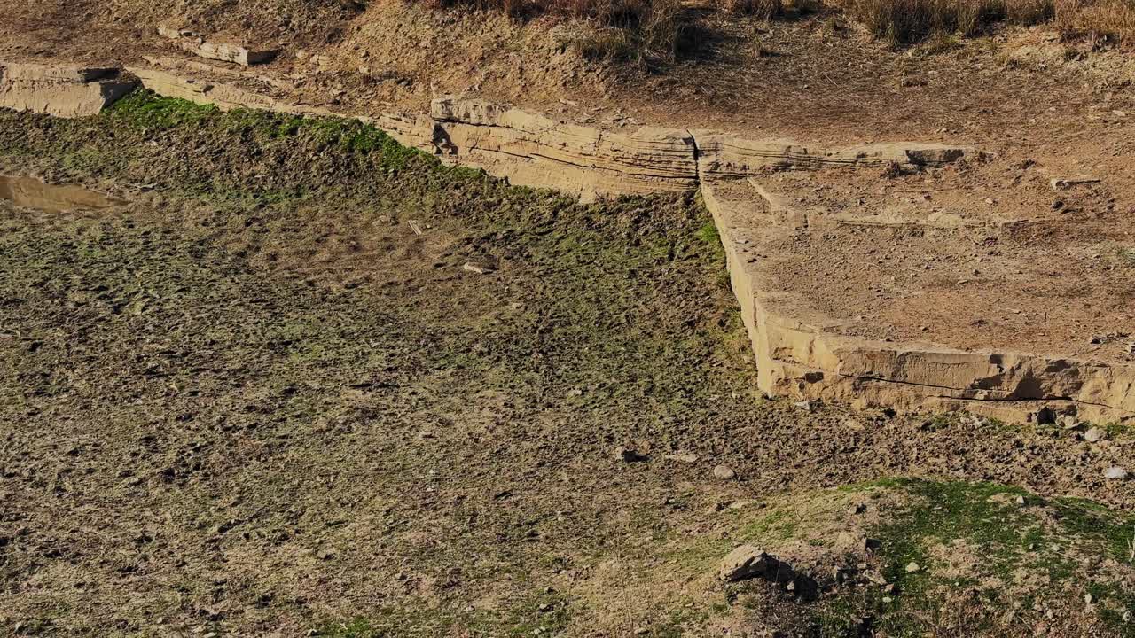 自然にできた池の溝によって露出した風化した石の層。乾燥した土の質感と近くに育つ新鮮な緑の小麦の作物で、土壌侵食のパターンを示しています。