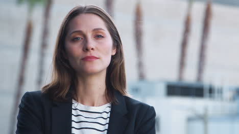 portrait of serious businesswoman standing outside office building with city skyline in background