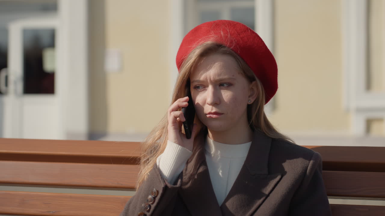 Giovane donna bianca su una panchina controlla il telefono con espressione preoccupata, indossa un berretto rosso e un cappotto di lana, sotto la calda luce solare. Sfondo di una facciata cittadina pallida, scorre i messaggi e poi risponde a una chiamata. Una tranquilla scena su una panchina del parco.