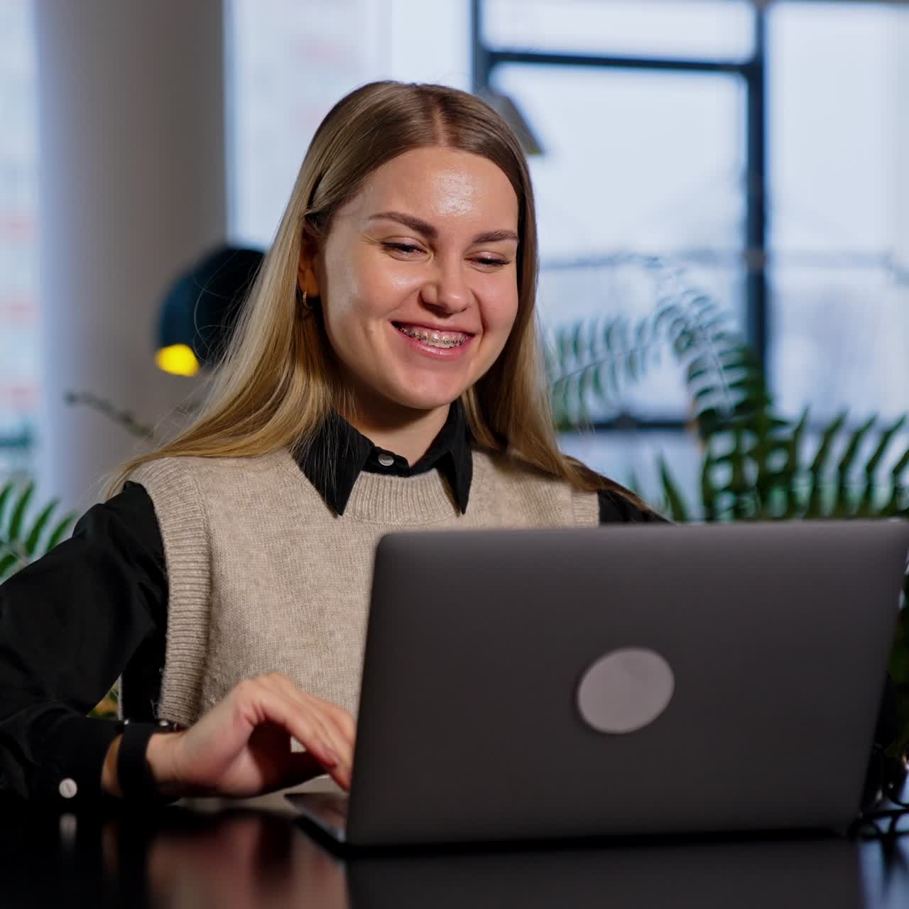 Resilient smiling young woman working on her computer. Happy lady is satisfied with some result and makes a success gesture. Blurred backdrop