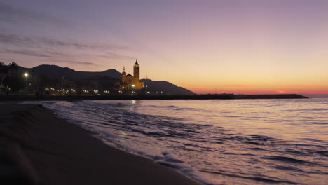 coastal-sunrise-skyline-is-reflected-in-sea-waves-washing-ashore,-silhouette-of-church-stands-against-backdrop-of-majestic-mountain-range