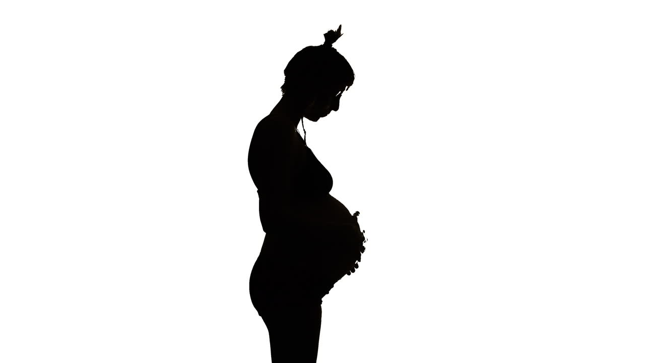 SILHOUETTE - A heavily pregnant woman looks down and strokes her belly