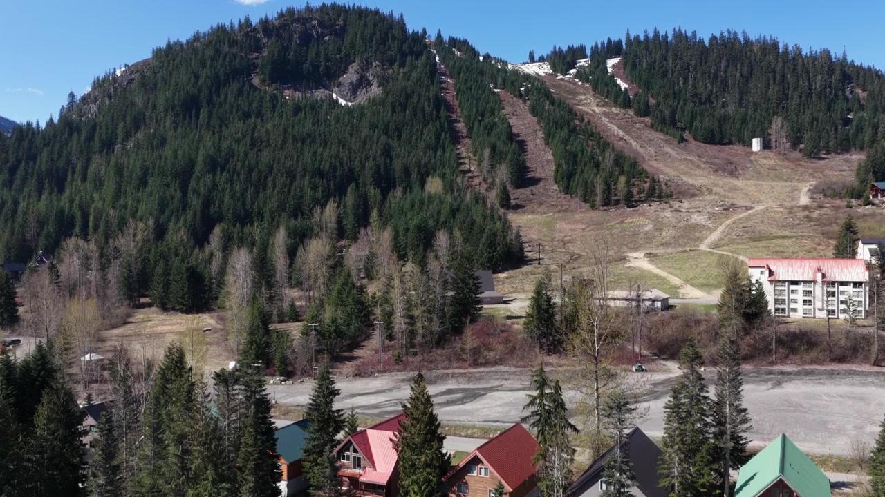 Fjellandsbyen Snoqualmie Pass med trehytter og koier under bare skiløyper, flekkvis snø på skogkledde skråninger og nakne vårtrær under lys blå himmel. Washington, USA. Flyfoto.