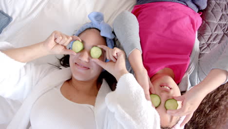 madre y hija bi-raciales felices acostadas en la cama con rebanadas de pepino en los ojos